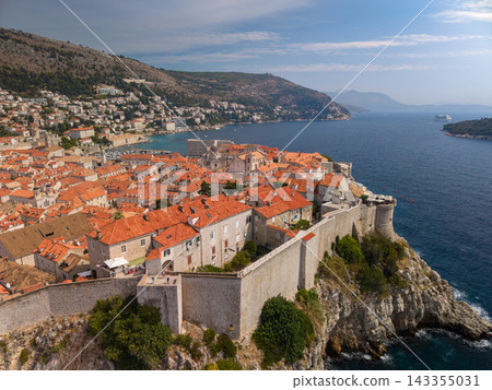 Dubrovnik Croatia fills coastal view with terracotta rooftops, stone walls and blue Adriatic Sea. Historic old town sits beneath wooded hills, with cruise ship visible offshore in clear daylight. Dubrovnik Croatia fills coastal view with terracotta rooftops, stone walls and blue Adriatic Sea. Historic old town sits beneath wooded hills, with cruise ship visible offshore in clear daylight. 143355031