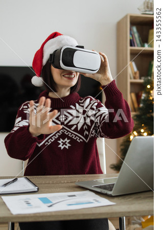 A Caucasian woman wearing a Santa hat and festive sweater uses a virtual reality headset while sitting at a desk with a laptop and papers 143355562