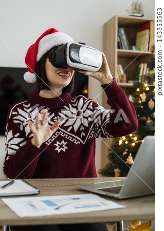 A Caucasian woman wearing a Santa hat and festive sweater uses a virtual reality headset while sitting at a desk with a laptop and Christmas tree in the background A Caucasian woman wearing a Santa hat and festive sweater uses a virtual reality headset while sitting at a desk with a laptop and Christmas tree in the background 143355563