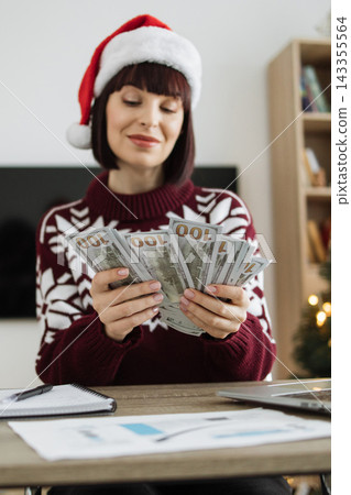 A Caucasian woman wearing a festive sweater and Santa hat counts a fan of US dollar bills at a desk with a laptop and papers 143355564