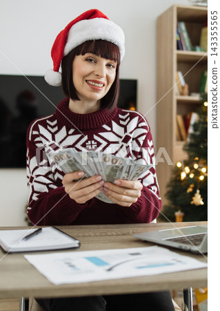 A smiling Caucasian woman wearing a festive sweater and Santa hat counts a fan of US dollar bills at a desk with a Christmas tree in the background A smiling Caucasian woman wearing a festive sweater and Santa hat counts a fan of US dollar bills at a desk with a Christmas tree in the background 143355565