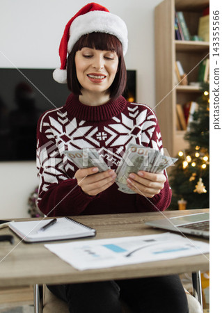 A woman wearing a Santa hat and festive sweater counts US dollar bills while sitting at a desk with a laptop and papers A woman wearing a Santa hat and festive sweater counts US dollar bills while sitting at a desk with a laptop and papers 143355566