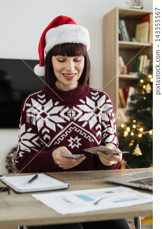 A woman wearing a Santa hat and festive sweater counts US dollar bills at a desk with a laptop and Christmas tree in the background A woman wearing a Santa hat and festive sweater counts US dollar bills at a desk with a laptop and Christmas tree in the background 143355567