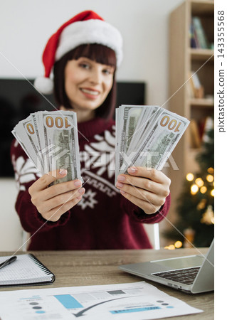A smiling Caucasian woman wearing a Santa hat and a festive sweater holds out stacks of US dollar bills, suggesting holiday bonuses or earnings A smiling Caucasian woman wearing a Santa hat and a festive sweater holds out stacks of US dollar bills, suggesting holiday bonuses or earnings 143355568