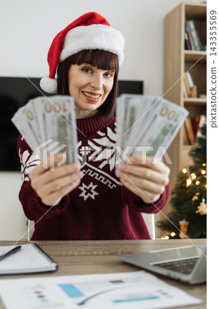 A smiling woman wearing a Santa hat and a festive sweater holds out US dollar bills, suggesting holiday bonuses or earnings A smiling woman wearing a Santa hat and a festive sweater holds out US dollar bills, suggesting holiday bonuses or earnings 143355569