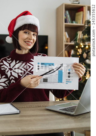 A smiling woman wearing a Santa hat and festive sweater presents a financial growth chart during the holiday season A smiling woman wearing a Santa hat and festive sweater presents a financial growth chart during the holiday season 143355612