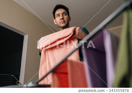 Shooting from below of modern man performing domestic tasks, folding clean laundry, contributing to household chores and gender equality in home life, demonstrating shared responsibilities. 143355878