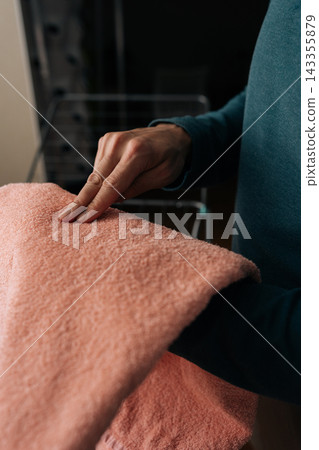 Cropped shot of person hand checking texture and softness of freshly laundered pink towel, hanging on indoor clothes drying rack, representing daily domestic tasks and fabric care. Cropped shot of person hand checking texture and softness of freshly laundered pink towel, hanging on indoor clothes drying rack, representing daily domestic tasks and fabric care. 143355879