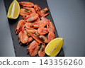 On a black stone board on a gray background are frozen shrimps, framed on both sides by lemon slices. Concept: symmetrical citrus frame, enhancing the bright emphasis on seafood. High quality photo 143356260