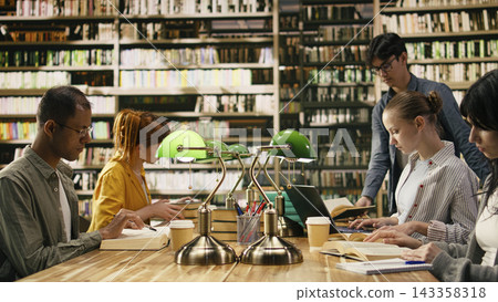 College students sitting in library reading books and using laptop, taking notes for coursework under green lamps. Focused academic study environment for learning, higher education. 143358318