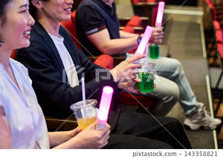 觀眾在劇院裡欣賞現場表演,手裡拿著螢光棒和飲料。 觀眾在劇院裡欣賞現場表演,手裡拿著螢光棒和飲料。 143358821