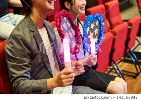 Young people watching the concert from the red seats, holding cheering goods. Young people watching the concert from the red seats, holding cheering goods. 143358842