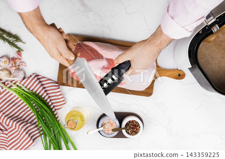 Man sharpening a kitchen knife with a sharpener to prepare raw meat. Male hands maintain blade edge over wooden cutting board with spices, garlic, green onion, and oil for cooking. Man sharpening a kitchen knife with a sharpener to prepare raw meat. Male hands maintain blade edge over wooden cutting board with spices, garlic, green onion, and oil for cooking. 143359225