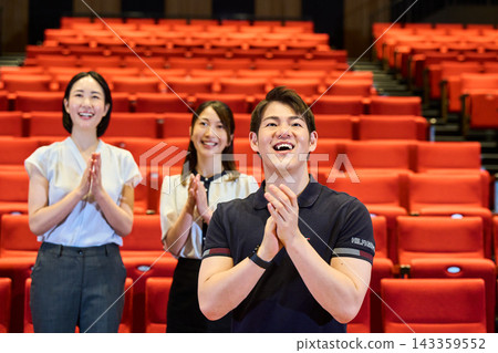 Men and women stand in the audience seats of the hall and applaud the stage in a show of admiration. Men and women stand in the audience seats of the hall and applaud the stage in a show of admiration. 143359552