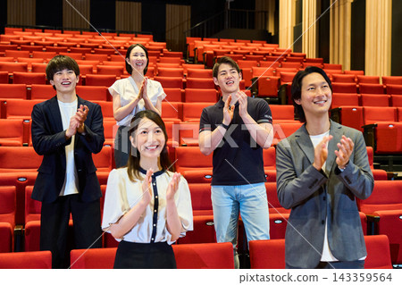 Men and women stand up in the hall's seating area and applaud towards the stage. Men and women stand up in the hall's seating area and applaud towards the stage. 143359564