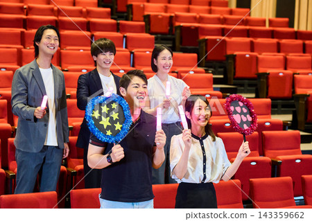 男男女女站在劇院裡,手持螢光棒和扇子,享受他們的扇子活動。 男男女女站在劇院裡,手持螢光棒和扇子,享受他們的扇子活動。 143359662