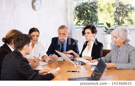 Concentrated young adult old office workers discussing ideas while gathering at table in meeting room Concentrated young adult old office workers discussing ideas while gathering at table in meeting room 143360635