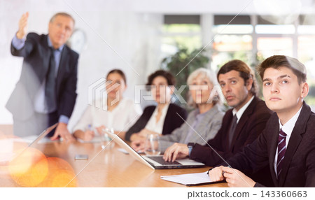 Office coworkers of different ages watching presentation while sitting at table with laptop and notes 143360663