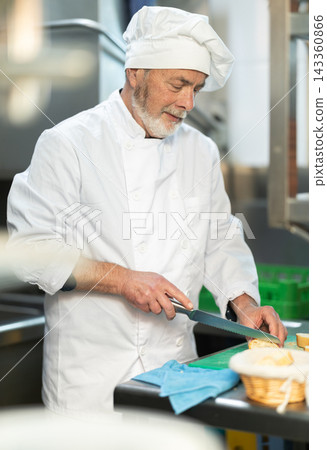 Senior man cook in kitchen finely slices food Senior man cook in kitchen finely slices food 143360866