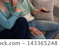 Young couple relaxing on sofa holding remote control while sitting close together at home 143365248