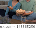 Young couple sharing popcorn bowl while holding remote control during movie night on sofa 143365250