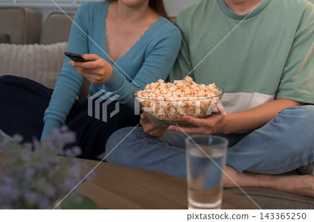 Young couple sharing popcorn bowl while holding remote control during movie night on sofa 143365250