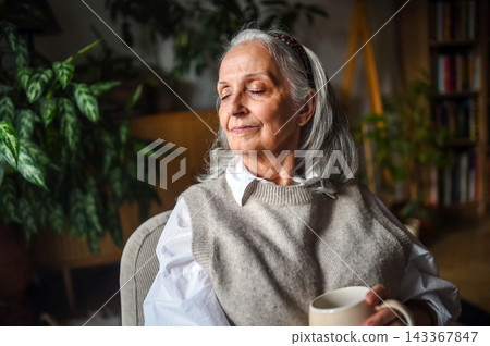 Peaceful senior woman relaxing with warm drink in cozy home. Peaceful senior woman relaxing with warm drink in cozy home. 143367847