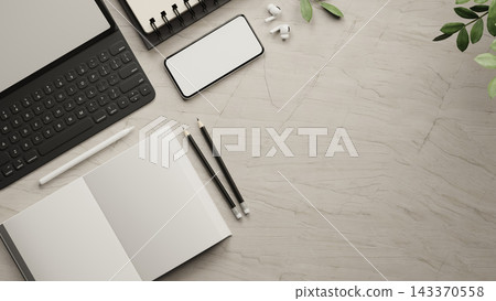 Top view of marble desk with tablet keyboard, blank smartphone, notebook, pencil, earbuds and leaves 143370558