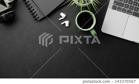 Top view of black desk with camera notebook earbuds green coffee cup laptop and plant top view Top view of black desk with camera notebook earbuds green coffee cup laptop and plant top view 143370567