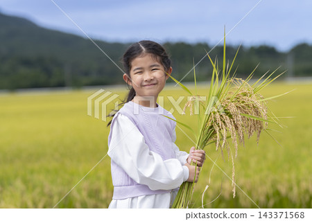一位笑容燦爛的6歲女孩手捧一捆米，站在稻田裡。 143371568