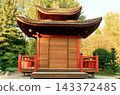 Traditional asian style wooden pagoda building with red railings and a multi-tiered tiled roof surrounded by green trees in a peaceful public garden park. 143372485