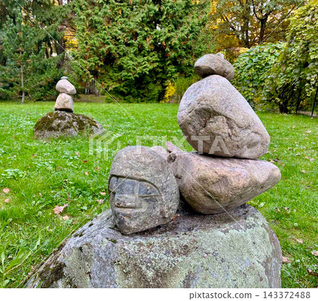 Stone balance and equilibrium concept on a green grass lawn with an ancient looking carved stone head face sculpture in a public park. 143372488