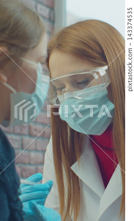 Vertical video. Professional doctor making injection to Caucasian child girl patient in mask at home during pandemic. Analysis. Vaccination concept. 143374535