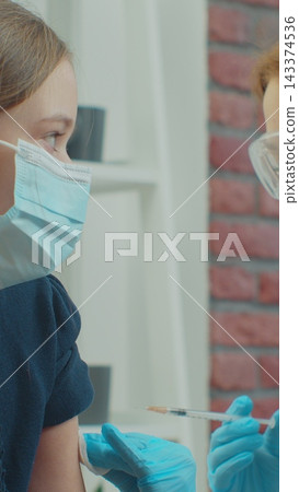 Vertical video. Female doctor wearing face mask injects a vaccine into the girl shoulder at home sitting on a couch. self isolation medical communication at home during pandemic. Vertical video. Female doctor wearing face mask injects a vaccine into the girl shoulder at home sitting on a couch. self isolation medical communication at home during pandemic. 143374536