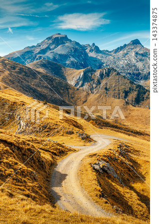 Winding dirt road through golden mountain slopes at Colle di Tenda pass with alpine peaks, France Winding dirt road through golden mountain slopes at Colle di Tenda pass with alpine peaks, France 143374875