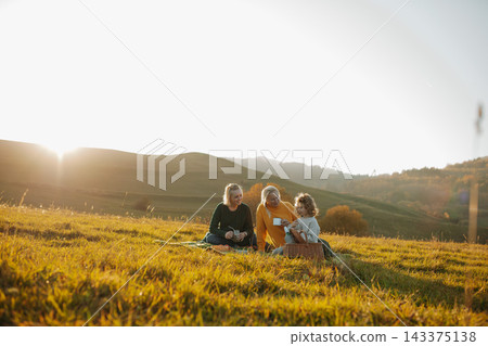 Three generations of women having a picnic on a green autumn meadow at sunset. Three generations of women having a picnic on a green autumn meadow at sunset. 143375138
