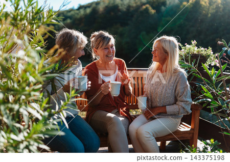 Senior female friends enjoying coffee together on cabin terrace getaway. 143375198