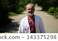 Elderly man wearing an embroidered shirt walking through rural road at sunny summer say. Senior male in traditional vyshyvanka going along country trail. Concept of national identity and patriotism 143375298