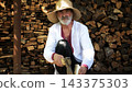 Older man wearing an embroidered shirt gently trims a piece of wood with axe against background of woodshed. Concept of craftsmanship, tradition and ukrainian heritage. Slow motion 143375303