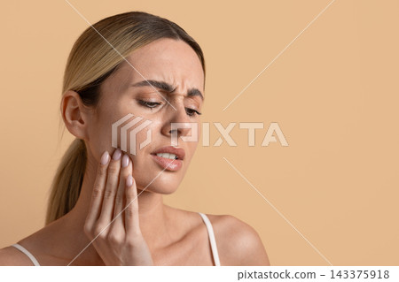 Young woman holding her jaw in discomfort against beige studio background 143375918