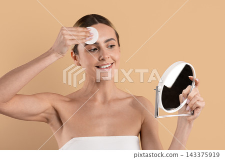 Smiling young woman wiping forehead with cotton pad while looking into mirror Smiling young woman wiping forehead with cotton pad while looking into mirror 143375919