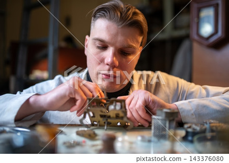 Young craftsman assembling intricate brass clock movement on workshop bench Young craftsman assembling intricate brass clock movement on workshop bench 143376080