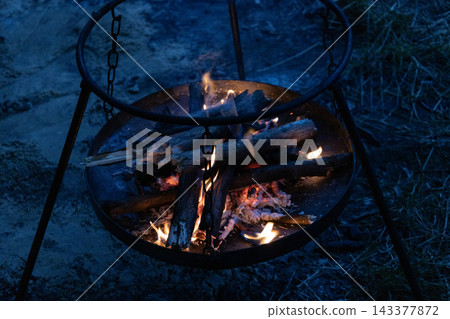 Metal campfire ring with a burning wood fire at night. Glowing flames and embers warm the dark outdoor setup. Portable tripod stand holds a round iron hearth during a cozy wilderness camping trip. Metal campfire ring with a burning wood fire at night. Glowing flames and embers warm the dark outdoor setup. Portable tripod stand holds a round iron hearth during a cozy wilderness camping trip. 143377872