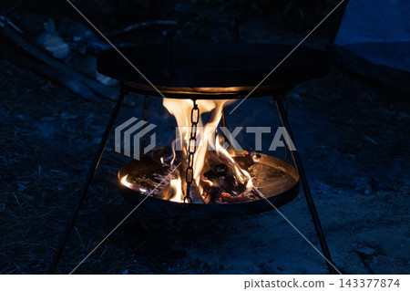 Campfire glowing brightly under a black metal frying pan at night. Hot flames burn logs in a portable outdoor stove stand. Warm light illuminates the dark forest setting during a cozy wilderness Campfire glowing brightly under a black metal frying pan at night. Hot flames burn logs in a portable outdoor stove stand. Warm light illuminates the dark forest setting during a cozy wilderness 143377874