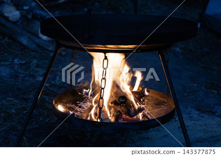 Campfire glowing brightly under a black metal frying pan at night. Hot flames burn logs in a portable outdoor stove stand. Warm light illuminates the dark forest setting during a cozy wilderness Campfire glowing brightly under a black metal frying pan at night. Hot flames burn logs in a portable outdoor stove stand. Warm light illuminates the dark forest setting during a cozy wilderness 143377875