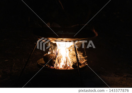 Campfire cooking set. A tripod holds a black metal pan over an open wood fire in the dark night. Glowing flames heat the cooking surface for preparing a hot meal outdoors in nature. Vertical tripod. Campfire cooking set. A tripod holds a black metal pan over an open wood fire in the dark night. Glowing flames heat the cooking surface for preparing a hot meal outdoors in nature. Vertical tripod. 143377876