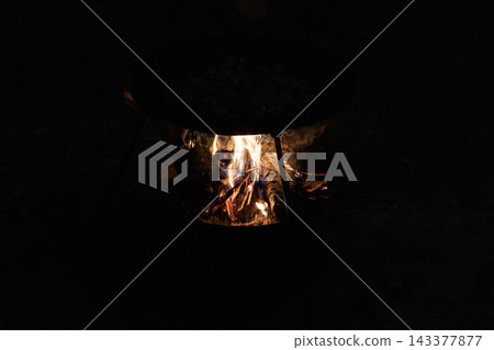 Campfire cooking set. A tripod holds a black metal pan over an open wood fire in the dark night. Glowing flames heat the cooking surface for preparing a hot meal outdoors in nature. Vertical tripod. Campfire cooking set. A tripod holds a black metal pan over an open wood fire in the dark night. Glowing flames heat the cooking surface for preparing a hot meal outdoors in nature. Vertical tripod. 143377877
