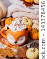 autumn background. Cute orange fox mug filled with hot chocolate topped with whipped cream, marshmallows and cinnamon, styled with white pumpkins, knitted blankets, autumn leaves and rustic fall 143378456
