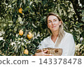 Smiling woman holding fresh lemons while sitting under a lemon tree in a sunny orchard 143378478