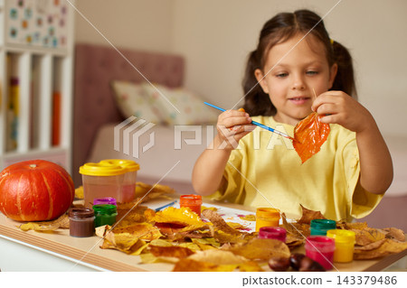 Young child creating autumn craft, painting a fall leaf with bright watercolors at a table with pumpkins and dry leaves Young child creating autumn craft, painting a fall leaf with bright watercolors at a table with pumpkins and dry leaves 143379486